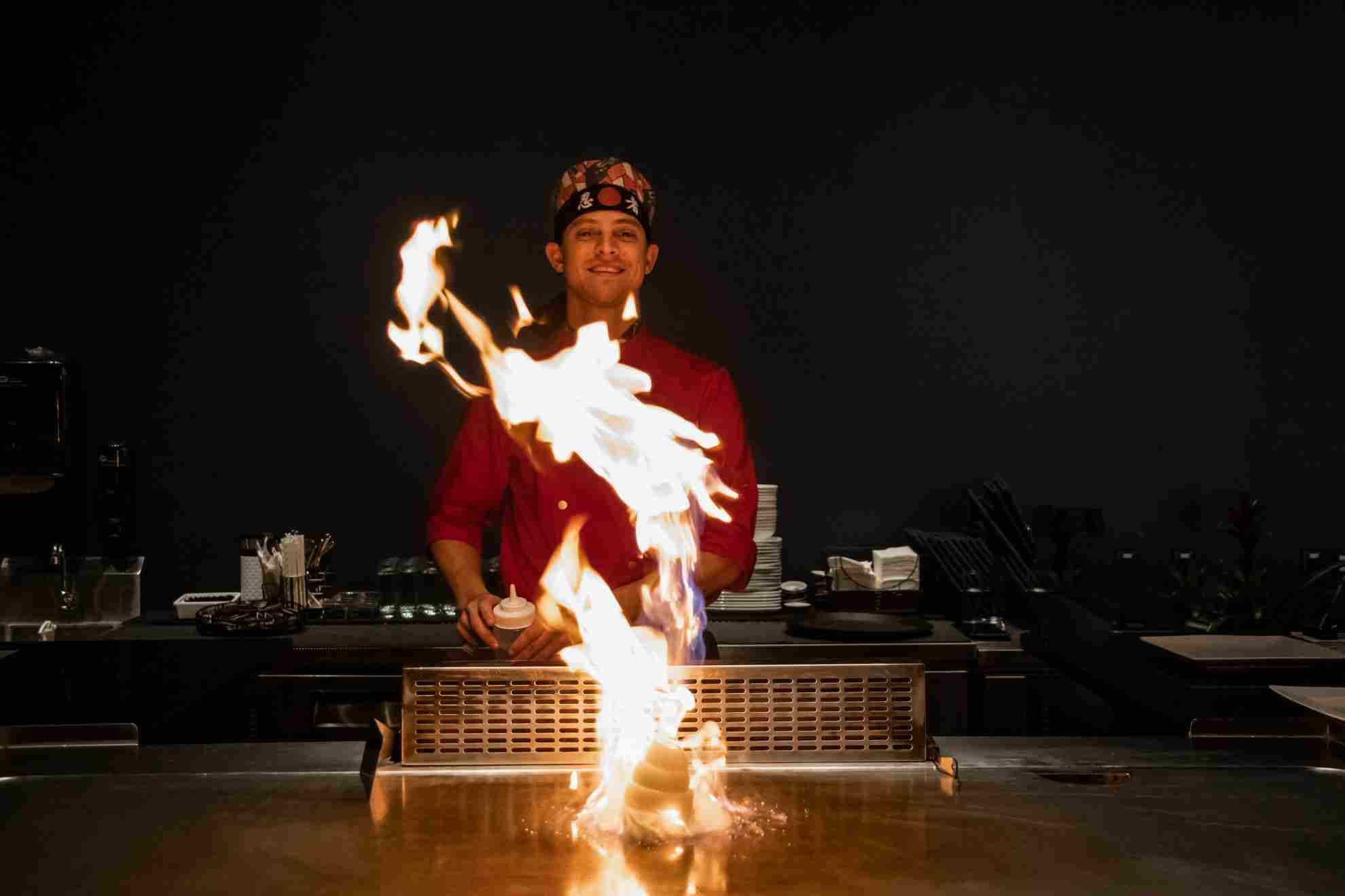 A Kasai teppanyaki chef smiling behind a column of flame rising from the grill