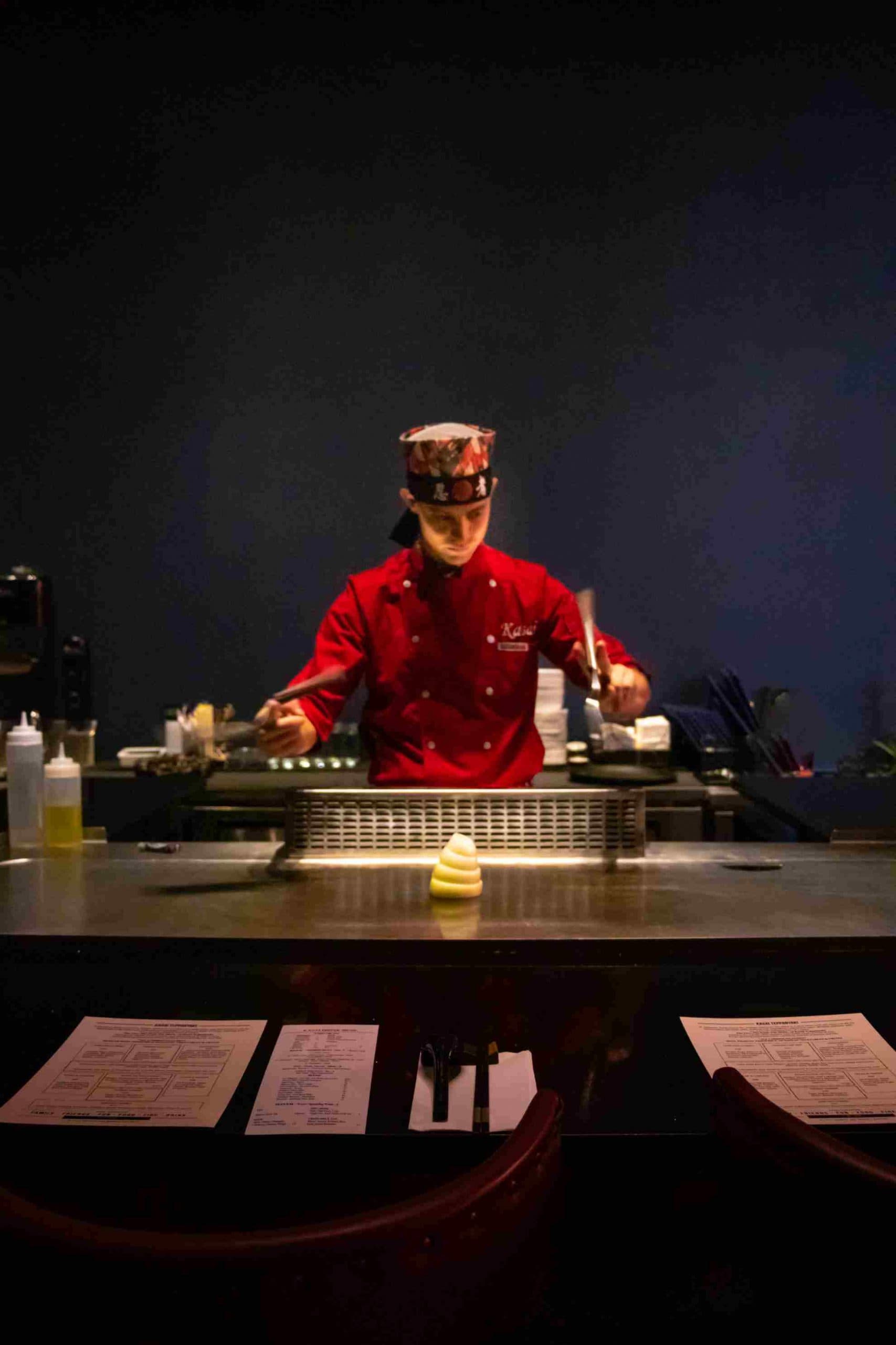 A Kasai chef in a red uniform working the teppanyaki grill under low light